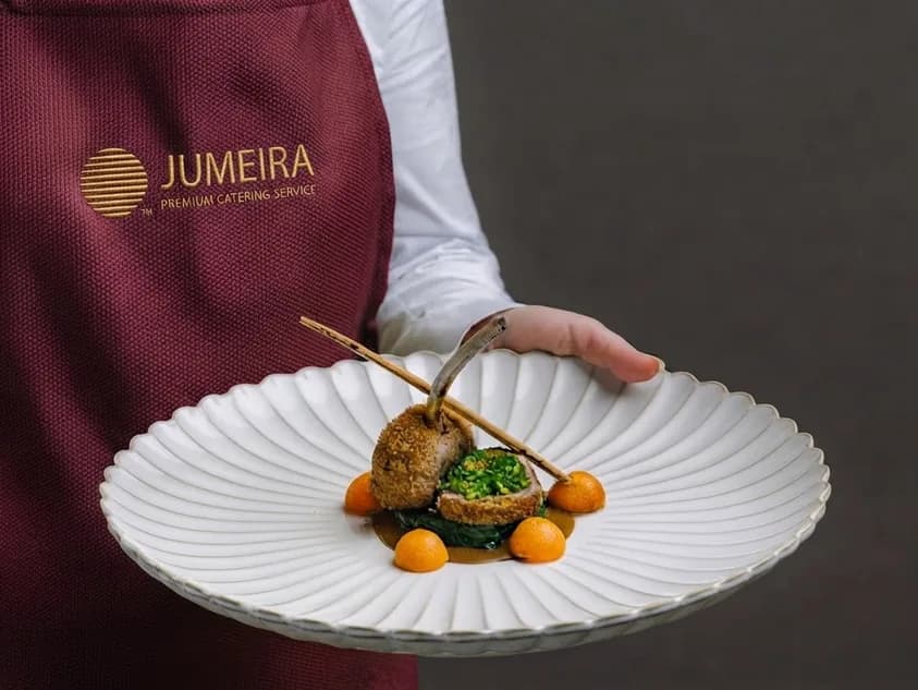A waiter in a Jumeira apron carrying a dish on a ribbed white plate