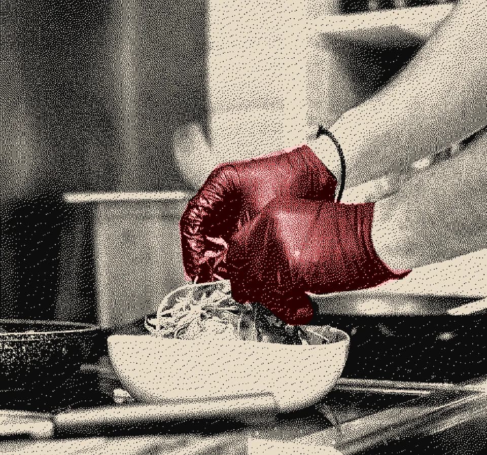 A gloved hand lifting a garnish over a white bowl in a working kitchen
